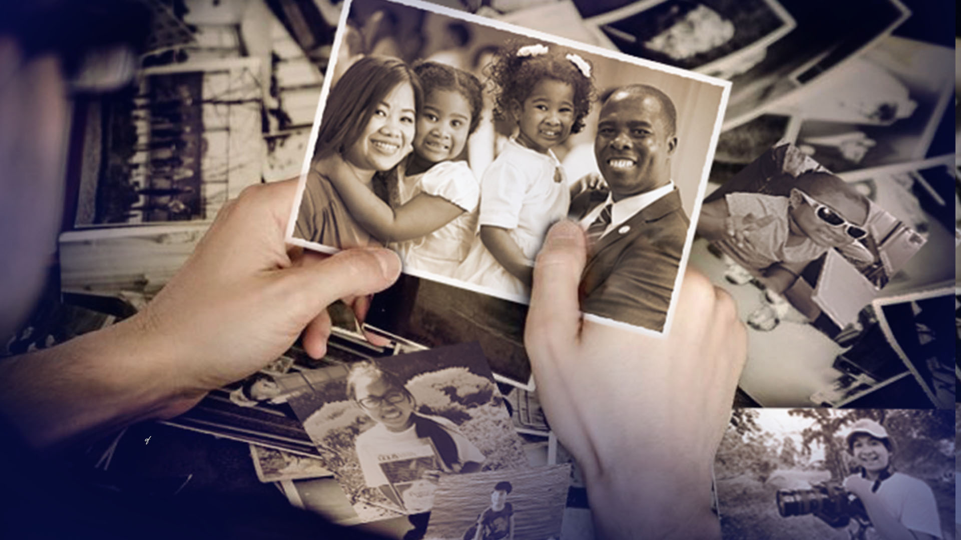 A pair of hands holds a single photo of a smiling family; on the table beneath is a pile of photos spread out.